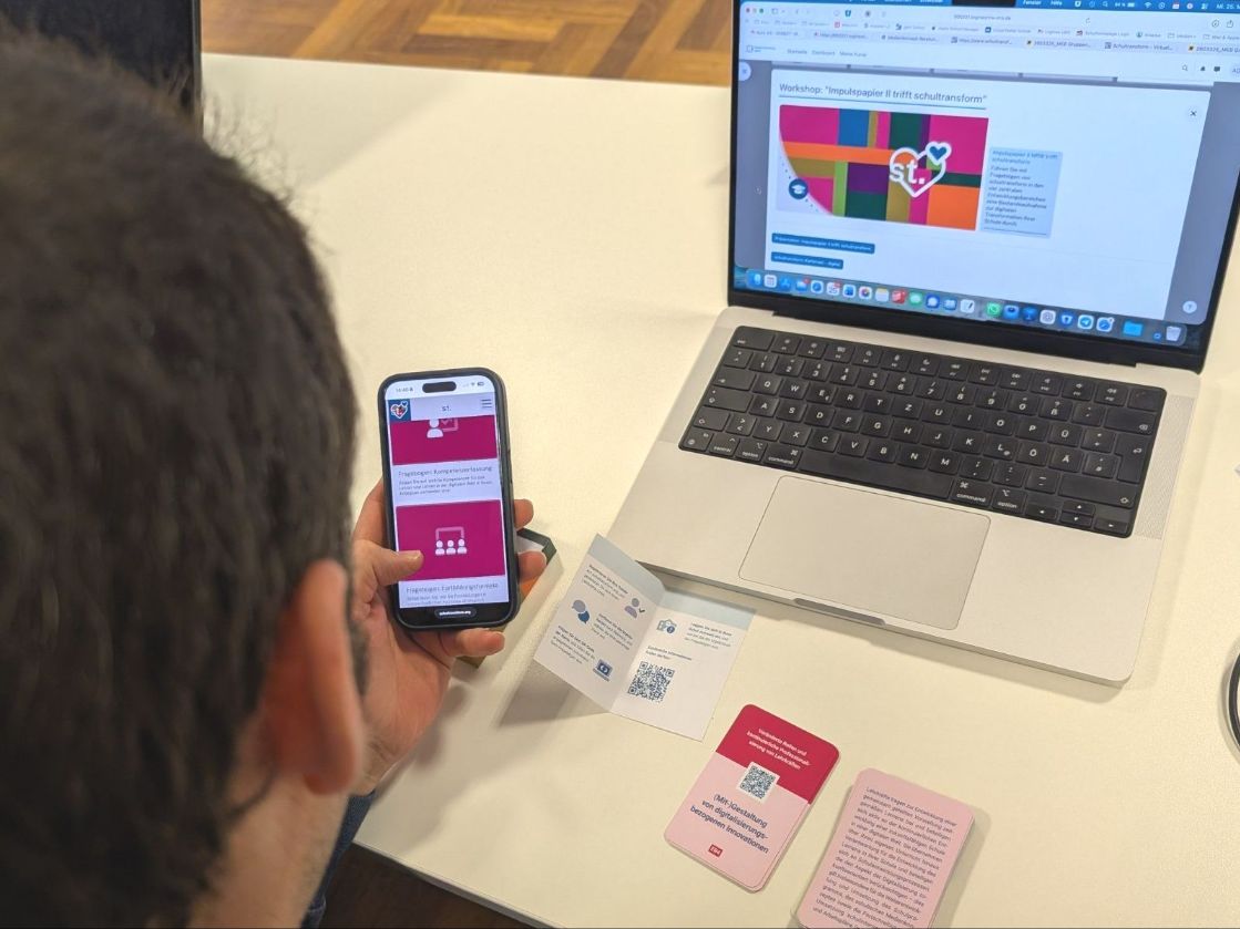 Ein Mann betrachtet die Plattform schultransform am Laptop und Smartphone. Das ergänzende analoge Kartenset liegt vor ihm auf dem Tisch. 