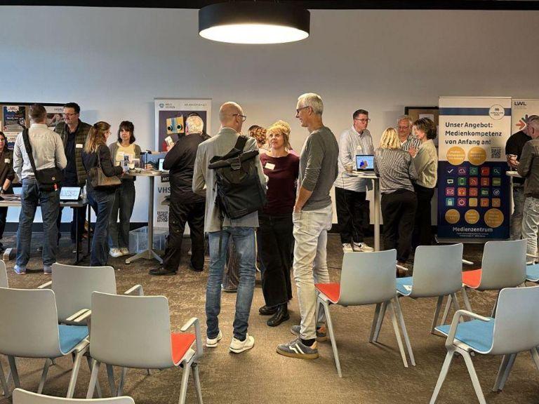 Menschen stehen in Gruppen zusammen und tauschen sich aus. Im Hintergrund stehen RollUps verschiedener Institutionen.	 Menschen stehen in Gruppen zusammen und tauschen sich aus. Im Hintergrund stehen RollUps verschiedener Institutionen.