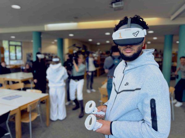 Ein männlicher Schüler trägt eine VR-Brille und hält Controller in seinen Händen. Im Hintergrund wird eine Schülerin durch ein Team des WDR via Kamera aufgenommen. Weitere Menschen bewegen sich durch den Klassenraum. Ein männlicher Schüler trägt eine VR-Brille und hält Controller in seinen Händen. Im Hintergrund wird eine Schülerin durch ein Team des WDR via Kamera aufgenommen. Weitere Menschen bewegen sich durch den Klassenraum.